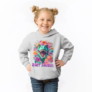 May include: A gray hooded sweatshirt with a colorful floral design featuring a dinosaur head with the text "Rory Saurus" printed on the front.