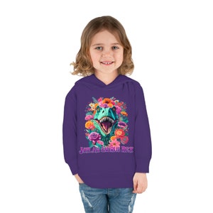 May include: Purple hooded sweatshirt with a colorful floral design and a dinosaur head in the center. The text "Jaylah Saurus Rex" is printed below the dinosaur.