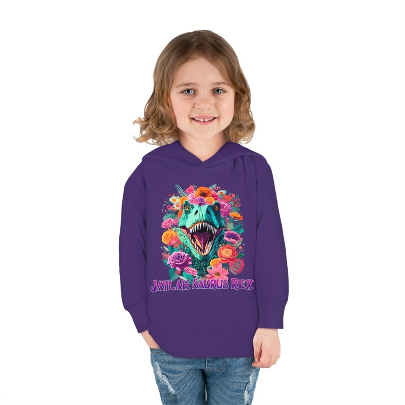 May include: Purple hooded sweatshirt with a colorful floral design and a dinosaur head in the center. The text "Jaylah Saurus Rex" is printed below the dinosaur.