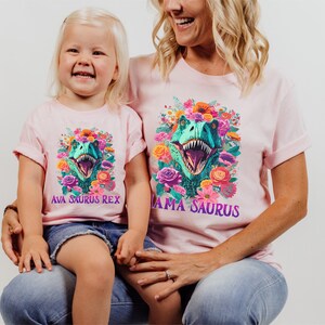 May include: A pink t-shirt with a floral design and a dinosaur head in the center. The text "Mama Saurus" is printed on the shirt. A child is wearing a pink t-shirt with a floral design and a dinosaur head in the center. The text "Ava Saurus Rex" is printed on the shirt.
