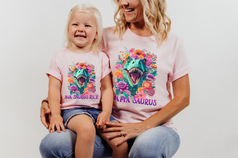 May include: A pink t-shirt with a floral design and a dinosaur head in the center. The text "Mama Saurus" is printed on the shirt. A child is wearing a pink t-shirt with a floral design and a dinosaur head in the center. The text "Ava Saurus Rex" is printed on the shirt.