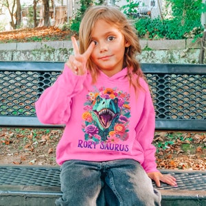 May include: A pink hoodie with a floral design and a dinosaur graphic. The dinosaur has a floral crown and the text "Rory Saurus" is printed below the graphic.