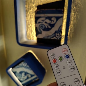 May include: Two blue-framed wall sconces with illuminated, patterned ceramic tile inserts. A remote control is in the foreground. The tile features a blue and white floral design. The sconces are mounted on a gold-colored wall.