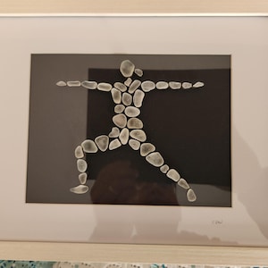 May include: Framed artwork featuring a yoga pose crafted from smooth, light-colored stones against a black background. The stones are arranged to depict a person in the Warrior II pose, set within a white mat and a light wood frame.