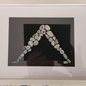 May include: Framed artwork featuring a black background with a white, arch-shaped design made of small, rounded stones. The artwork is encased in a white frame, creating a minimalist aesthetic. The piece is signed in the bottom right corner.
