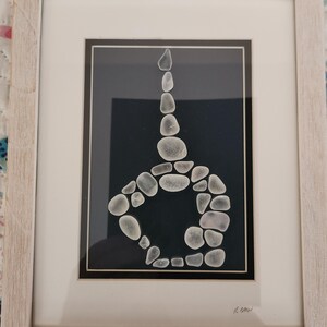 May include: Framed artwork featuring a design made of sea glass on a dark blue background. The sea glass is arranged in a stylized shape, with a light-colored wooden frame. The artwork is signed in the bottom right corner.