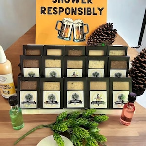 May include: A display of 12 different beer-themed soap bars with labels featuring various beer styles, such as Vanilla Porter, Citra IPA, and Cherry Lager. The display is on a wooden surface with a sign that reads "Shower Responsibly" and a beer-related definition.
