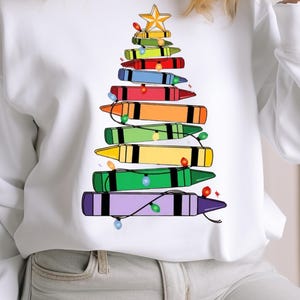 May include: White sweatshirt with a Christmas tree design made from crayons in various colours. The tree is topped with a yellow star and decorated with fairy lights. A festive holiday garment.