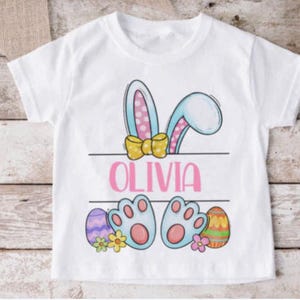 May include: White t-shirt with a colorful Easter bunny design. The bunny has pink and blue ears with white polka dots, a yellow bow, and blue feet. The shirt also has the name "Olivia" printed on it.