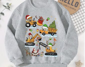 Puede incluir: Una sudadera gris con un diseño navideño con vehículos de construcción, Papá Noel y un árbol de Navidad. El texto "HELLO YOU LOVELY PEOPLE" está impreso en un posavasos de corcho en el fondo.