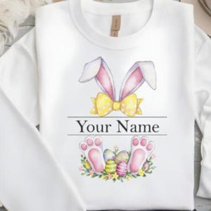 May include: White long-sleeved sweatshirt with a watercolor design of bunny ears, a yellow bow, and Easter eggs. The text "Your Name" is printed below the design.