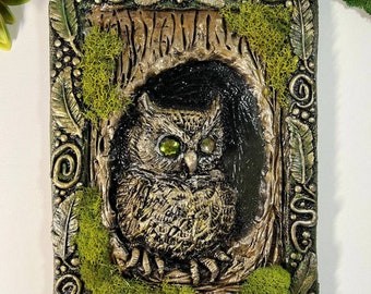 May include: A 3D wall art piece featuring a sculpted owl with green eyes, nestled in a tree trunk with moss and a decorative frame.