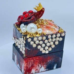 Pode incluir: Uma pequena caixa quadrada de resina vermelha, preta e branca com um acabamento brilhante. A caixa é decorada com uma figura de tartaruga vermelha que usa uma coroa dourada, glitter dourado e pérolas brancas. A caixa é perfeita para guardar pequenos objetos como joias ou bugigangas.