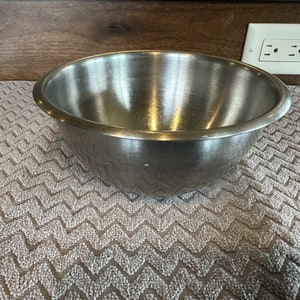 May include: A large, stainless steel mixing bowl sits on a beige, textured surface. The bowl is empty and has a shiny, reflective surface.