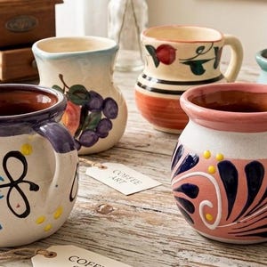 May include: A collection of five round ceramic mugs featuring various hand-painted designs, including floral patterns, fruit motifs, and abstract swirls in colors like blue, pink, green, and orange, with visible brown clay interiors and small paper tags.