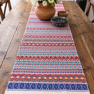 May include: A colorful table runner with a vibrant, geometric pattern in red, blue, yellow, and teal. The runner is placed on a wooden table and features a cream-colored fringe at the ends. A vase of white flowers and two decorative bowls sit on top.