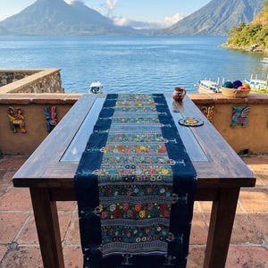 May include: A dark wood table with a glass top is adorned with a vibrant, embroidered table runner. The runner features a dark blue background with colorful floral patterns. The scene is set outdoors with a lake and mountains in the background.