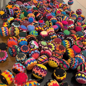 May include: A large collection of colorful, hand-crocheted hacky sacks. The hacky sacks feature a variety of vibrant colors and geometric patterns, including red, blue, yellow, green, and purple. The collection is displayed on a reflective surface.