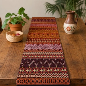 May include: A colorful woven table runner with a geometric pattern in shades of red, orange, and purple. The runner is displayed on a wooden table, alongside a small potted plant, a woven basket, and a decorative pitcher.