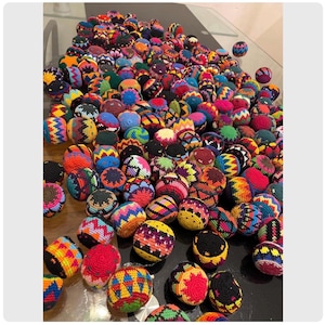 May include: A large collection of colorful, hand-crocheted balls. The balls feature a variety of vibrant colors and geometric patterns, including red, yellow, blue, and green. The balls are scattered across a reflective surface, creating a visually appealing display.