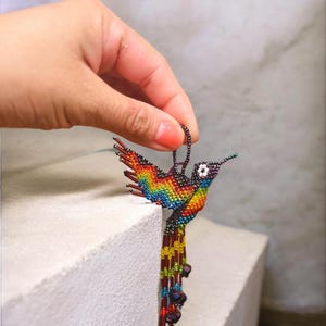 May include: A handcrafted hummingbird ornament made of colorful beads. The bird features a rainbow of colors on its wings and body, with a long beaded tail. The ornament is held by a hand, showcasing the intricate beadwork.
