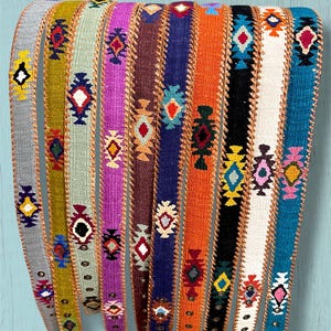 May include: A collection of ten woven belts featuring various solid base colors including gray, mustard, light green, purple, brown, navy, orange, black, white, and teal, each decorated with a repeating geometric pattern and stitched edges.