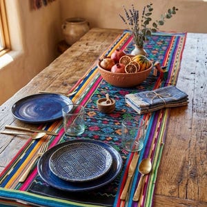 May include: A rustic wooden table is set with a vibrant, patterned runner. The table setting includes blue plates, clear glasses, gold-toned cutlery, and a terracotta bowl filled with fruit. A vase of lavender and folded napkins complete the scene.