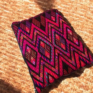 May include: A rectangular, zippered pouch with a vibrant, geometric pattern in shades of pink, red, purple, and blue. The design features zigzags and diamond shapes against a black background. The pouch is resting on a woven, tan surface.