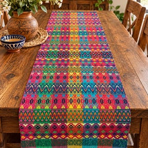 Puede incluir: Un camino de mesa rectangular con un patrón tejido vibrante y multicolor que presenta formas geométricas, zigzags y motivos florales en tonos rojo, azul, amarillo, verde y morado, acabado con pequeñas borlas coloridas en el borde inferior.