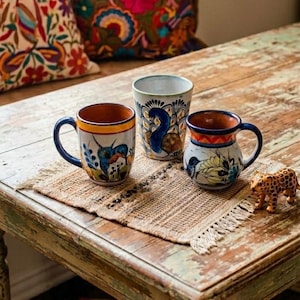 May include: Three ceramic mugs with colorful hand-painted designs sit on a woven placemat atop a rustic wooden table. The mugs feature blue, yellow, and orange accents, with images of birds and a peacock. A small leopard figurine is also on the table.