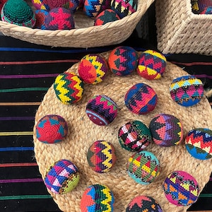 May include: A collection of colorful, hand-crocheted balls with geometric patterns. The balls are in various colors, including red, blue, yellow, and green, and are displayed in woven baskets and on a woven mat. The balls appear to be handmade and are likely used for juggling or play.