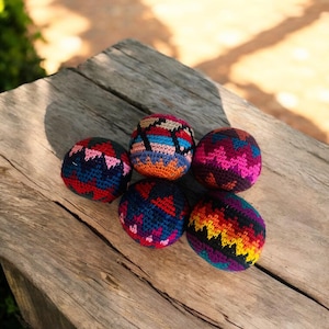 Puede incluir: Seis bolas coloridas, tejidas a mano, descansan sobre una superficie de madera envejecida. Las bolas presentan patrones vibrantes en tonos de rojo, azul, rosa, naranja y amarillo. Las bolas son un artículo lúdico y decorativo.
