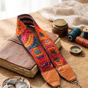 May include: A camera strap featuring vibrant, multi-colored floral embroidery in shades of orange, red, blue, and green. The strap includes tan leather end tabs with metal swivel hooks for attachment, providing a secure and decorative accessory.