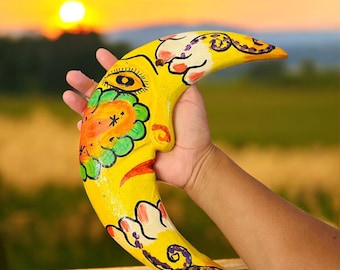 May include: A hand-painted, yellow crescent moon sculpture with a whimsical design. The moon features a face with an eye, floral accents, and orange and green details. The artwork is held against a sunset backdrop.