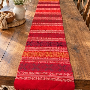 Puede incluir: Un camino de mesa rectangular largo con una base de color rojo vibrante y bandas horizontales de intrincados patrones geométricos en tonos naranja, amarillo, rojo oscuro y negro, terminado con un borde de flecos en el extremo inferior.