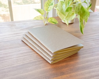 May include: Four blank, beige notebooks with rounded corners are stacked on a wooden surface. The notebooks are neatly arranged, with the top one slightly open. A potted plant with green and yellow leaves is in the background.