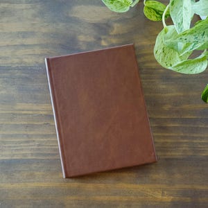 以下が含まれることがあります： 茶色の革張りの閉じたノートが、濃い木製の表面に置かれています。 ノートの表紙は滑らかで長方形です。 上部右隅には、緑と白の斑入りの葉があり、自然な要素が加えられています。