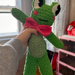 May include: A handmade green crochet frog plushie with a pink bow tie. The frog has white and black eyes and pink cheek accents. The plushie is made of a textured yarn and has outstretched arms.