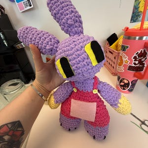 May include: A handmade crochet bunny doll in shades of purple, pink, and yellow. The bunny has large ears, black eyes with yellow accents, and wears pink overalls with a felt pocket. The doll is a soft, plush toy.