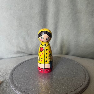 May include: A hand-painted wooden doll wearing a yellow raincoat with red polka dots, a red heart on the chest, and red boots. The doll is holding a black umbrella.