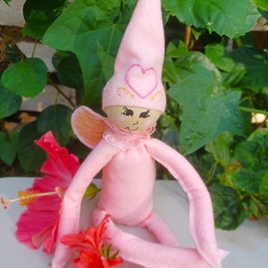 Puede incluir: Una muñeca de hada de fieltro rosa con un corazón rosa en su sombrero y una flor rosa en su mano. Está sentada sobre una superficie blanca con hojas verdes en el fondo.