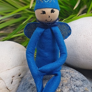 Könnte beinhalten: Eine blaue Filzpuppe mit einer spitzen Mütze, die ein gesticktes Herz und goldene Akzente aufweist. Die Puppe hat einen runden Holzkopf mit einem handgezeichneten Gesicht, dunkelblaue Flügel mit goldenem Glitzer und lange, schlanke Gliedmaßen.