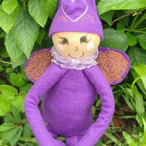 May include: A purple felt doll with a pointed hat featuring an embroidered white heart. The doll has a light-colored wooden face with painted facial features, a white ruffled collar, and purple wings with gold-colored speckles on the back.