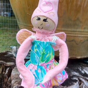 May include: A handmade fairy doll with a pink pointed hat and matching arms and legs. The doll has a wooden face with painted features and wears a colorful dress with a floral pattern. The fairy has small, tan wings.