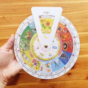 Könnte beinhalten: Ein weißes, kreisförmiges Kalenderrad mit einem farbenfrohen Design, das die Monate des Jahres und die Jahreszeiten darstellt. Das Rad ist in 12 Abschnitte unterteilt, die jeweils einen Monat repräsentieren. Die Monate sind in Deutsch beschriftet, die Jahreszeiten in Deutsch und Englisch. Das Rad ist auf August eingestellt, der in Deutsch mit "Sommer" beschriftet ist.