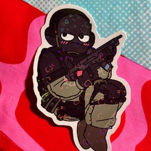 May include: A sticker featuring a cartoon soldier in tactical gear, kneeling and holding a rifle. The soldier is black with pink blush marks and hearts. The sticker has a holographic effect and is set against a red, pink, and blue patterned background.