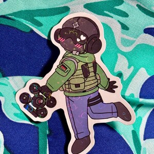 May include: A sticker featuring a cartoon character in a military-style outfit, including a helmet, vest, and pants. The character is holding a device with multiple circular components. The sticker has a white border and is set against a patterned background.