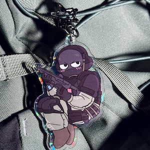 May include: A clear acrylic keychain featuring a cartoon soldier in a crouching position, holding a rifle. The soldier is wearing a black helmet, purple uniform, and tan pants. The keychain has a silver clasp attached to a green backpack.