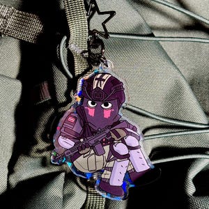 May include: A clear acrylic keychain with a cartoon soldier illustration. The soldier wears a helmet and tactical gear, including a rifle. The design features purple, grey, and black colours. The keychain is attached to a black star-shaped clasp.