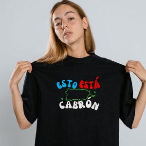 May include: A black t-shirt with a white graphic print of the text "ESTO ESTA CABRON" in a colorful, cartoon font. The graphic also includes a green outline of the island of Puerto Rico.